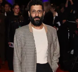 Adeel Akhtar, dressed in formal attire, walking on the red carpet with photographers in the background.