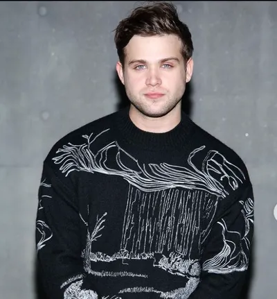 Leo Woodall in a sweater stands in front of a wall.